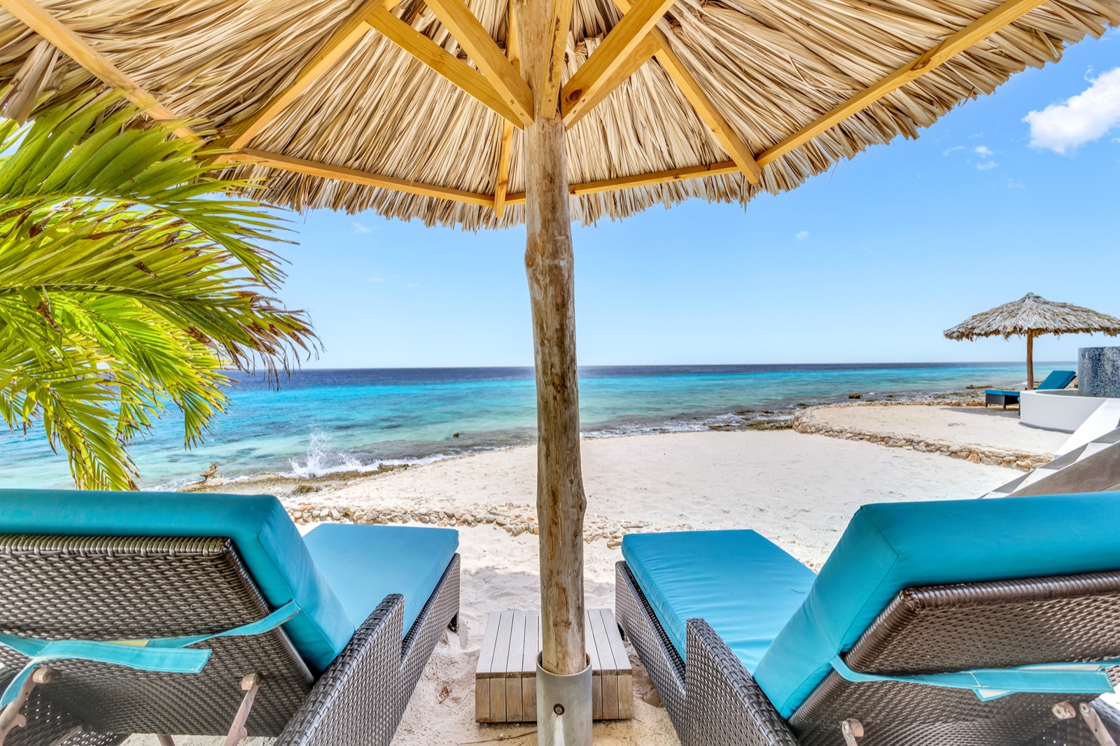 Luxury Bonaire villa with private beach access, featuring two cushioned lounge chairs under a thatched palapa umbrella looking out over the calm turquoise ocean.